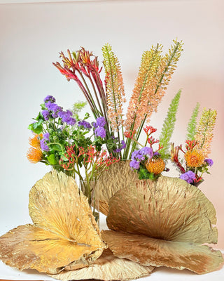 Composizione con fiori arancioni, viola e color pesca, verde ornamentale e grandi foglie dorate su fondo chiaro.